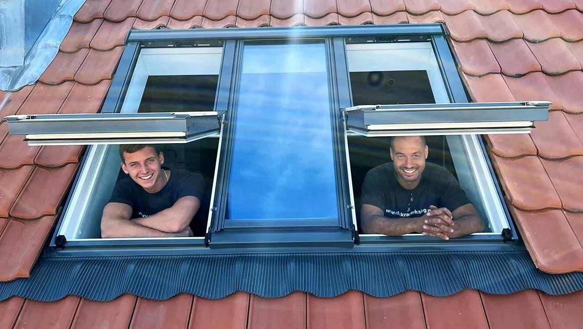Monteurs van Dakraamkoning kijken glimlachend uit twee geopende dakramen in een pannendak.
