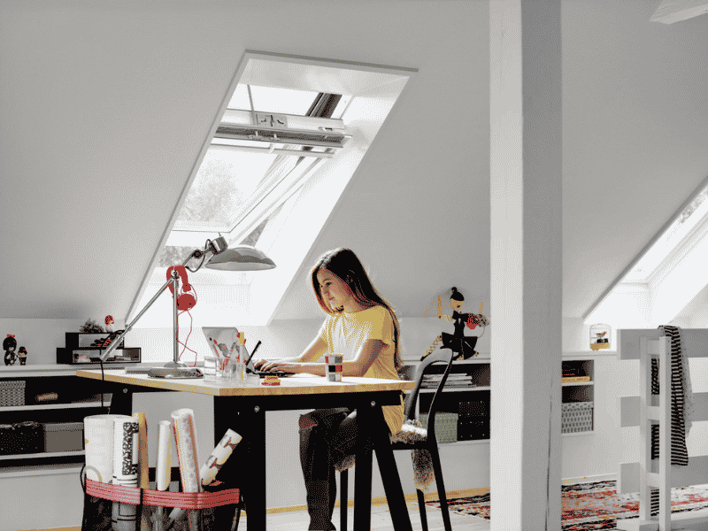 Zolderkamer met een elektrisch VELUX dakraam boven een bureau waar iemand werkt